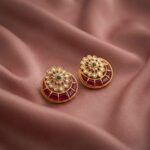 Antique gold-tone kundan style stud earrings with clear-stone flower centre, green accent and ruby-coloured crescent on dusty pink fabric