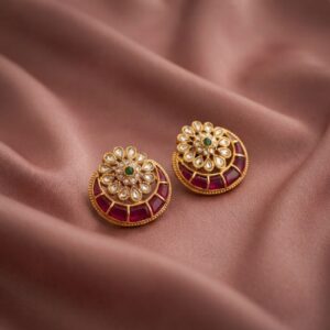 Antique gold-tone kundan style stud earrings with clear-stone flower centre, green accent and ruby-coloured crescent on dusty pink fabric