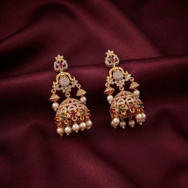 Gold-tone cubic zirconia jhumka earrings with ruby-coloured and green stones, side bells and faux pearl drops on burgundy fabric