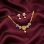 Gold-tone dainty necklace set with Lakshmi coin pendant, cubic zirconia flower medallions, faux pearl drop and matching stud earrings on burgundy fabric