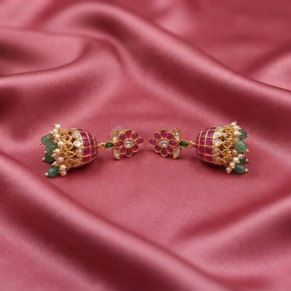 Antique gold-tone ruby red jhumka earrings with floral stud tops, lattice bells, green beads and faux pearl drops on dusty rose fabric