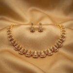 Gold-tone openwork wedding guest necklace set with cubic zirconia, ruby-coloured accents and matching triangular drop earrings on gold fabric