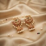 Antique gold-tone chandbali earrings with crescent frame, jhumka drop, ruby-coloured and green stones and faux pearls on champagne fabric
