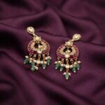 Gold-tone Indian chandelier earrings with ruby-coloured panels, clear teardrop stones, green bead fringe and faux pearl drops on plum fabric
