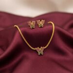 Gold-tone butterfly necklace set with pink and green stone-set butterfly pendant, woven chain and matching butterfly stud earrings on burgundy fabric