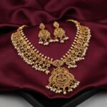 Antique gold-tone Hyderabadi necklace set with wide collar, ruby-coloured and green stones, faux pearl drops, ornate pendant and matching chandelier earrings on burgundy fabric