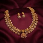 Antique gold-tone Nagas necklace set with Lakshmi motifs, ruby-coloured floral medallions, faux pearl drops and matching stud earrings on burgundy fabric
