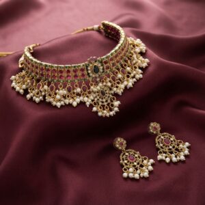 Kundan-style choker necklace set with ruby-coloured stones, pearl-effect drops and matching chandelier earrings on maroon fabric