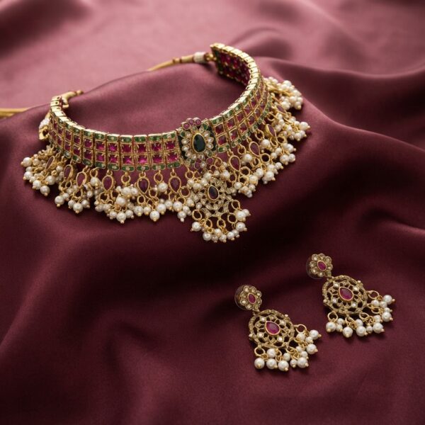 Kundan-style choker necklace set with ruby-coloured stones, pearl-effect drops and matching chandelier earrings on maroon fabric