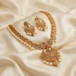 Matte gold-tone chandbali necklace set with crescent pendant, ruby-coloured and green stones, faux pearl drops and matching chandbali earrings on cream fabric