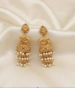 Antique gold-tone temple jhumka earrings with Lakshmi stud tops, cow motif, three-tier bells and faux pearl drops on cream fabric