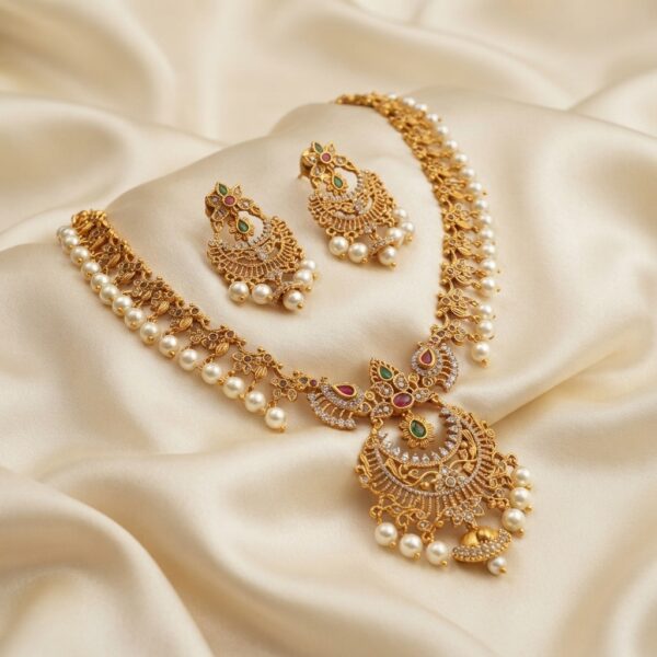 Matte gold-tone chandbali necklace set with crescent pendant, ruby-coloured and green stones, faux pearl drops and matching chandbali earrings on cream fabric