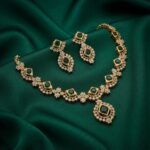 Gold-tone green stone necklace set with square-cut green stones, cubic zirconia halos, centre pendant and matching drop earrings on green fabric