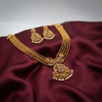 Antique gold-tone multi-strand bead temple jewellery necklace set with ornate pendant, pearl-effect drops and matching chandelier earrings on maroon fabric