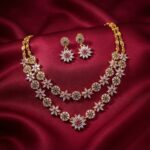 Gold-tone layered flower necklace set with two strands of cubic zirconia flowers, ruby-coloured and green centres and matching drop earrings on burgundy fabric