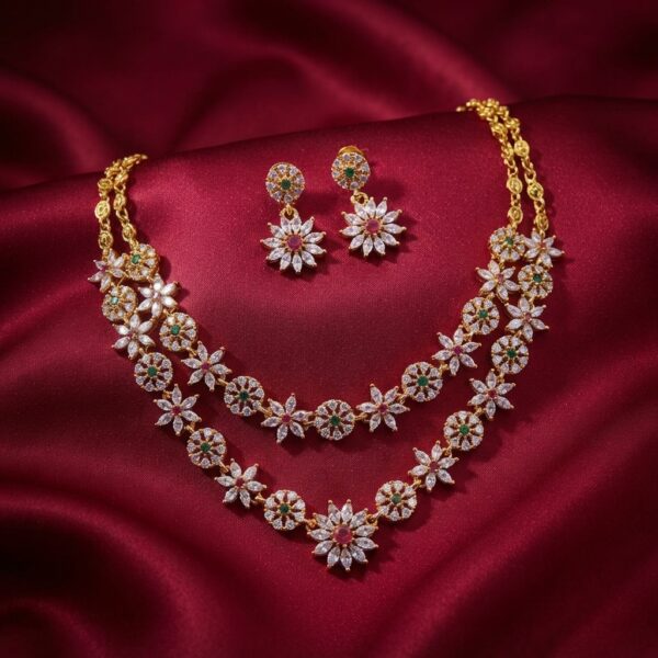 Gold-tone layered flower necklace set with two strands of cubic zirconia flowers, ruby-coloured and green centres and matching drop earrings on burgundy fabric