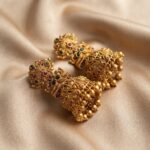 Antique gold-tone tiered jhumka earrings with textured scrollwork, ruby-coloured and green stones and gold bead drops on beige fabric